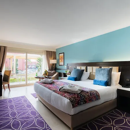 Savoy Le Grand Hotel Marrakech Marrakesh Buitenkant foto The photo shows a well-decorated hotel room featuring a large bed with colorful bedding and decorative pillows. The room has a light blue wall and a modern design with elegant furnishings. To the left, there are two windows with curtains, allowing na